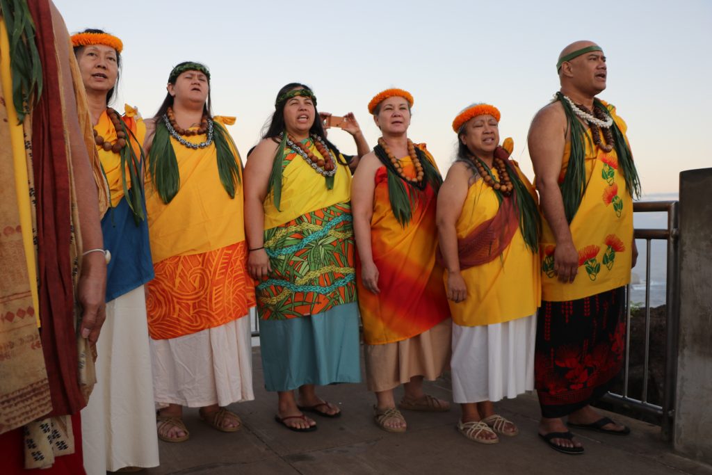 Seven cultural practitioners in Native Hawaiian dress stand side by side chanting an 'oli on the platform at the top of Makapu'u at sunrise.