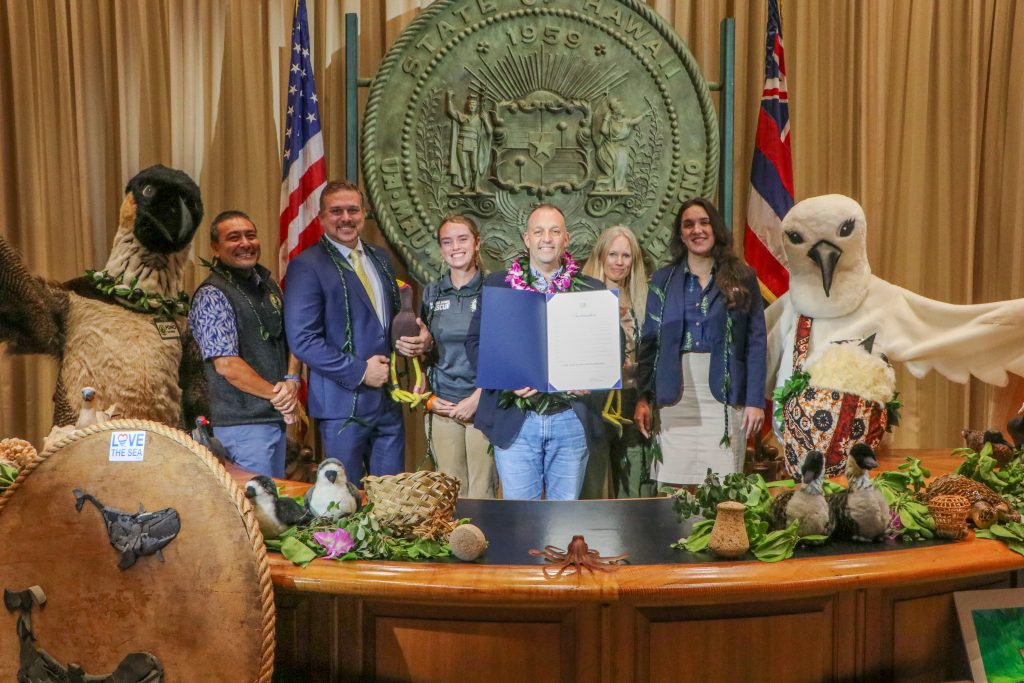 Five people posing with Governor Josh Green, who is holding the proclamation for "Year of Our Coastal Kuleana" in his office. People in a nene costume and a white tern costume stand on either side of the group.